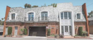 Image is of a grey and white shingled building with balconies on the top florr, and brick exterior on the bottom floor. 